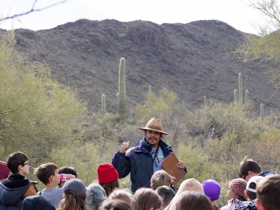 A man in a blue rain coat wearing a woven hat stands in the desert in front of a group of grade school children as he points behind him at the desert landscape.
