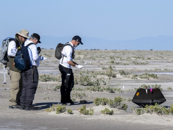three people approaching a tire-sized capsule on a desert landscape