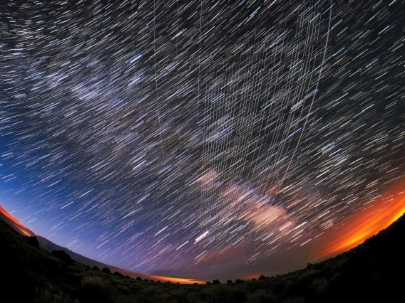 a fisheye image of a starry sky over a forest with white streaks through the sky